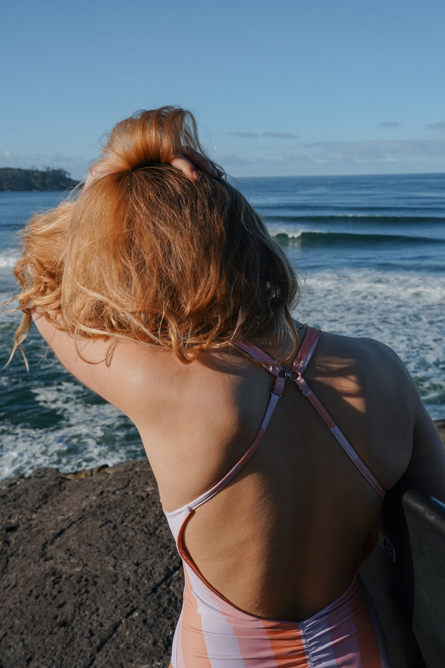 Back view of a striped one piece swimsuit with open back design in a coastal summer setting